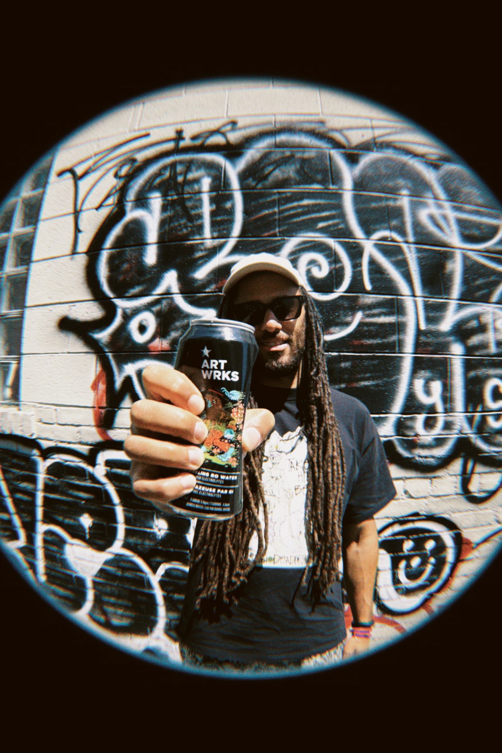 A man holds a black ART WRKS can toward the fisheye lens against a painted brick wall.