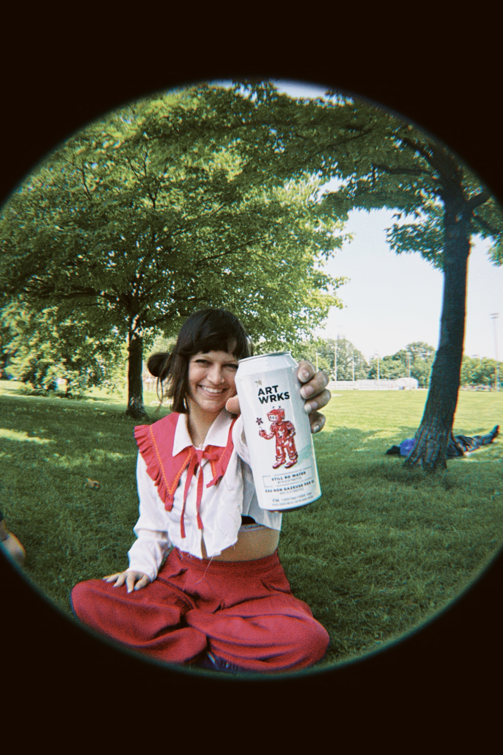 A woman in red and white sits on the grass, holding an ART WRKS can toward the fisheye lens.