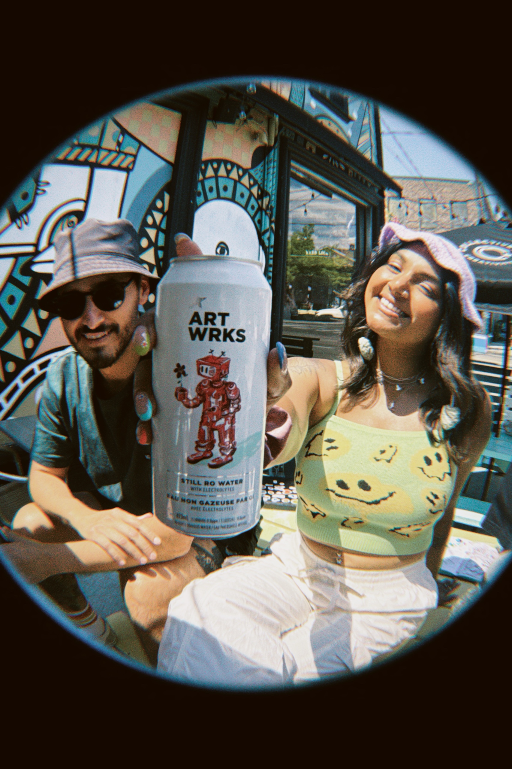 Two people outside a mural-covered storefront, holding an ART WRKS can toward the fisheye lens.