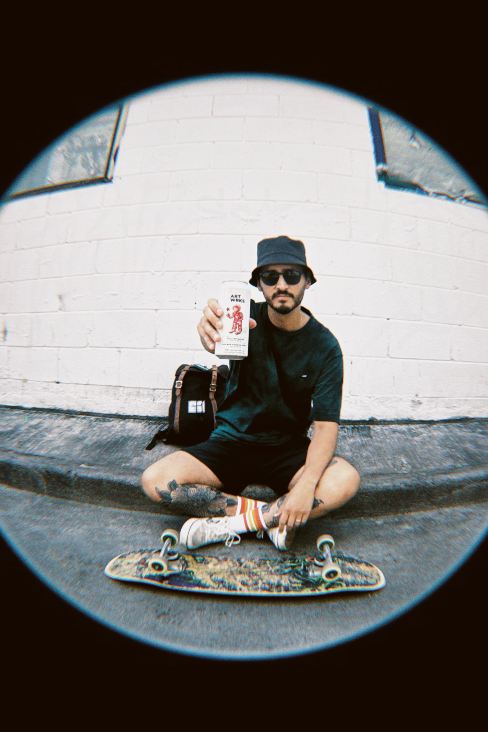 Fisheye portrait of a man sitting on a skateboard and holding an ART WRKS can.