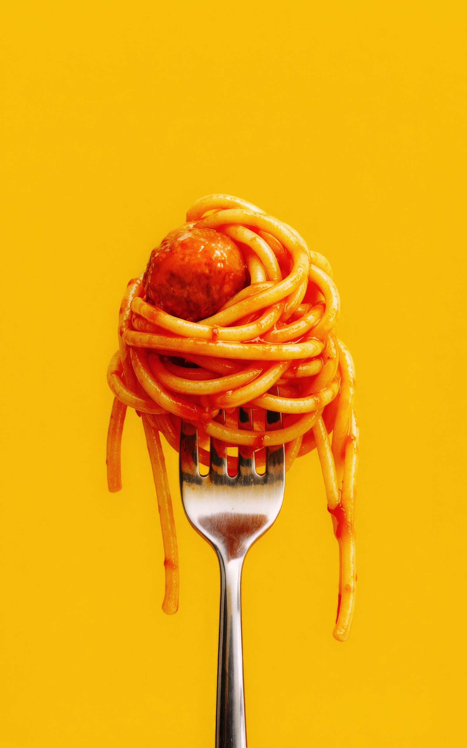 Spaghetti and a meatball wrapped around a fork against a bright yellow background.