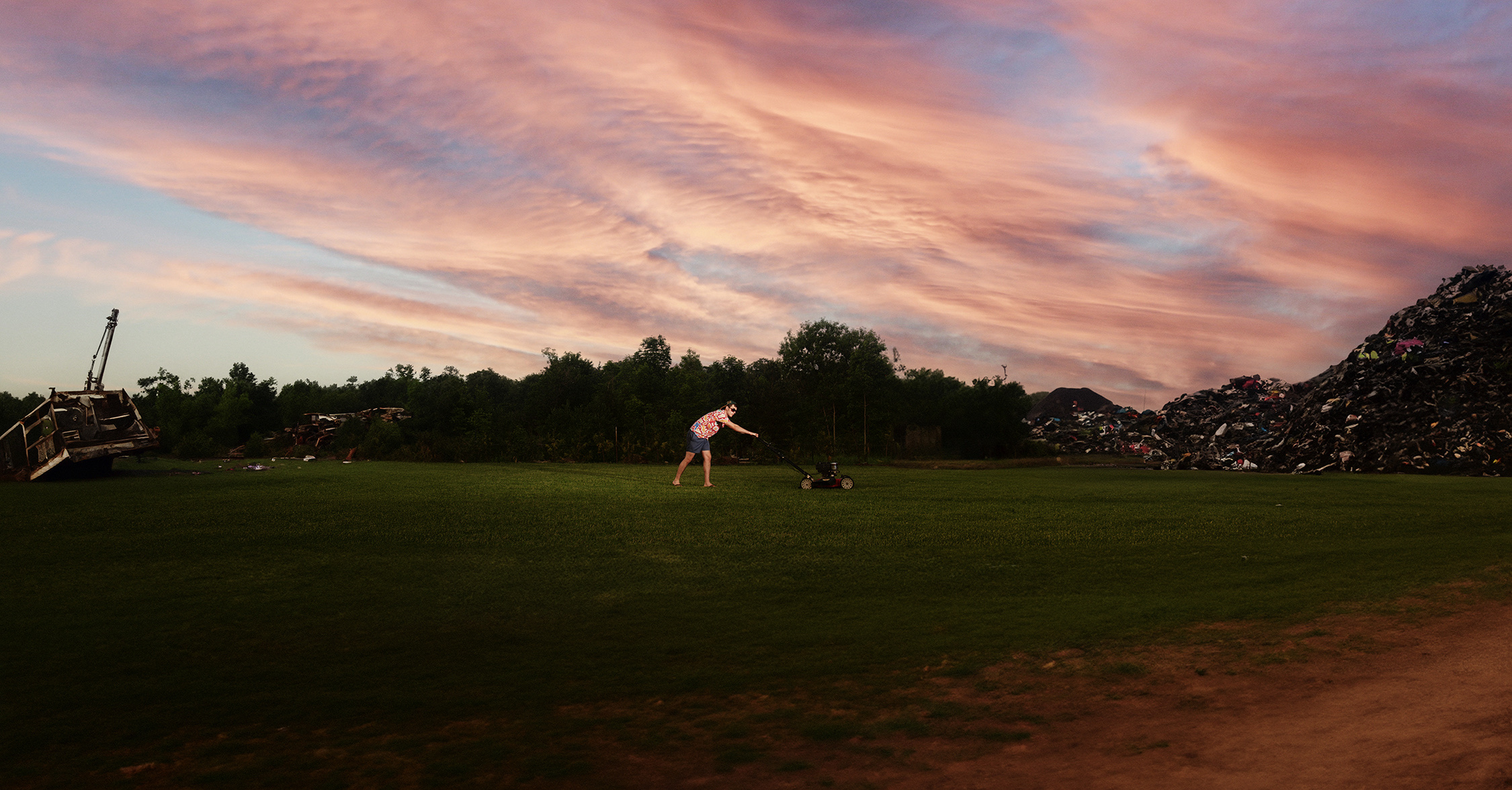 A man mows a lawn between scrap heaps beneath a sweeping pink sunset sky.