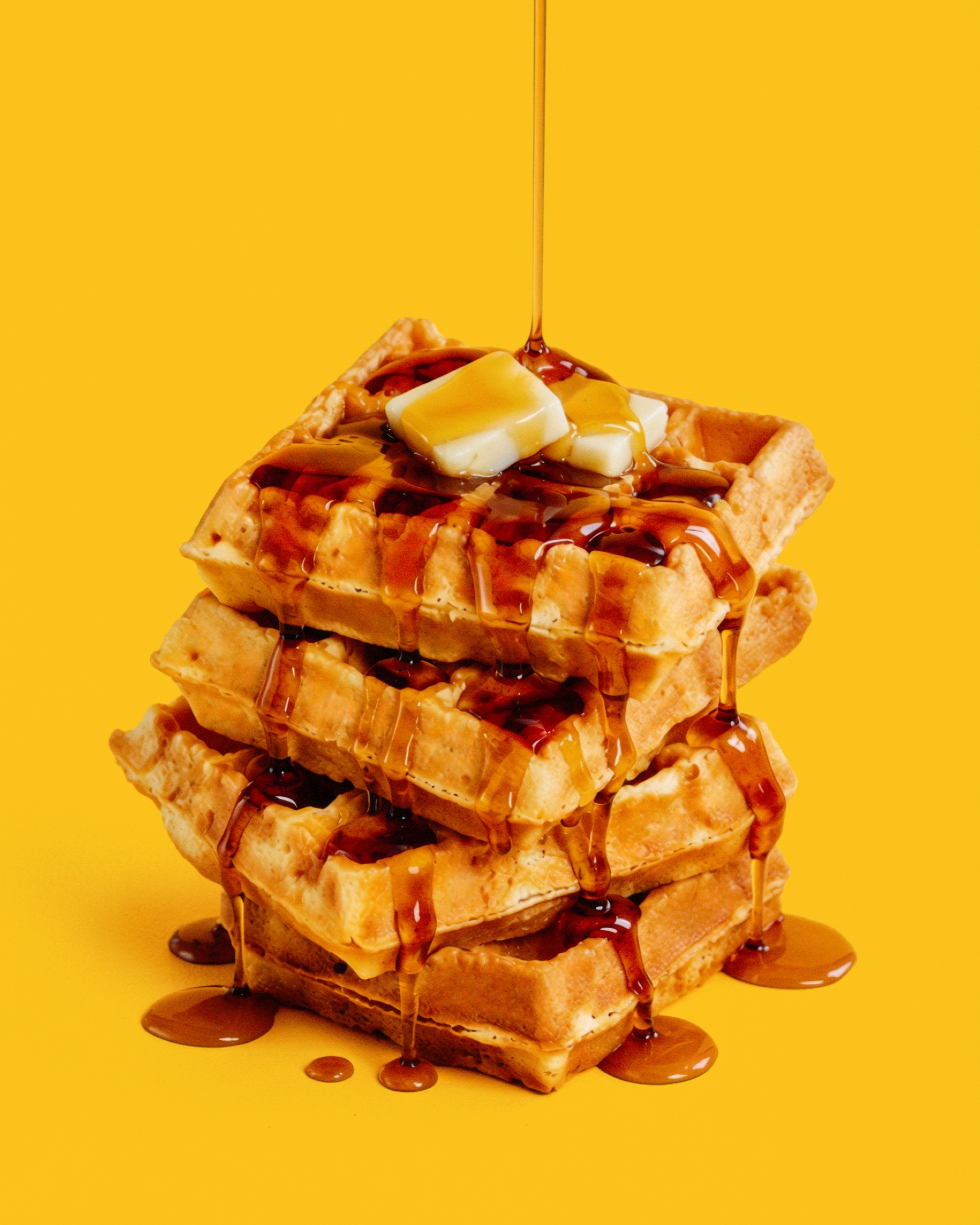 Syrup pours over a stack of waffles topped with butter against a yellow background.