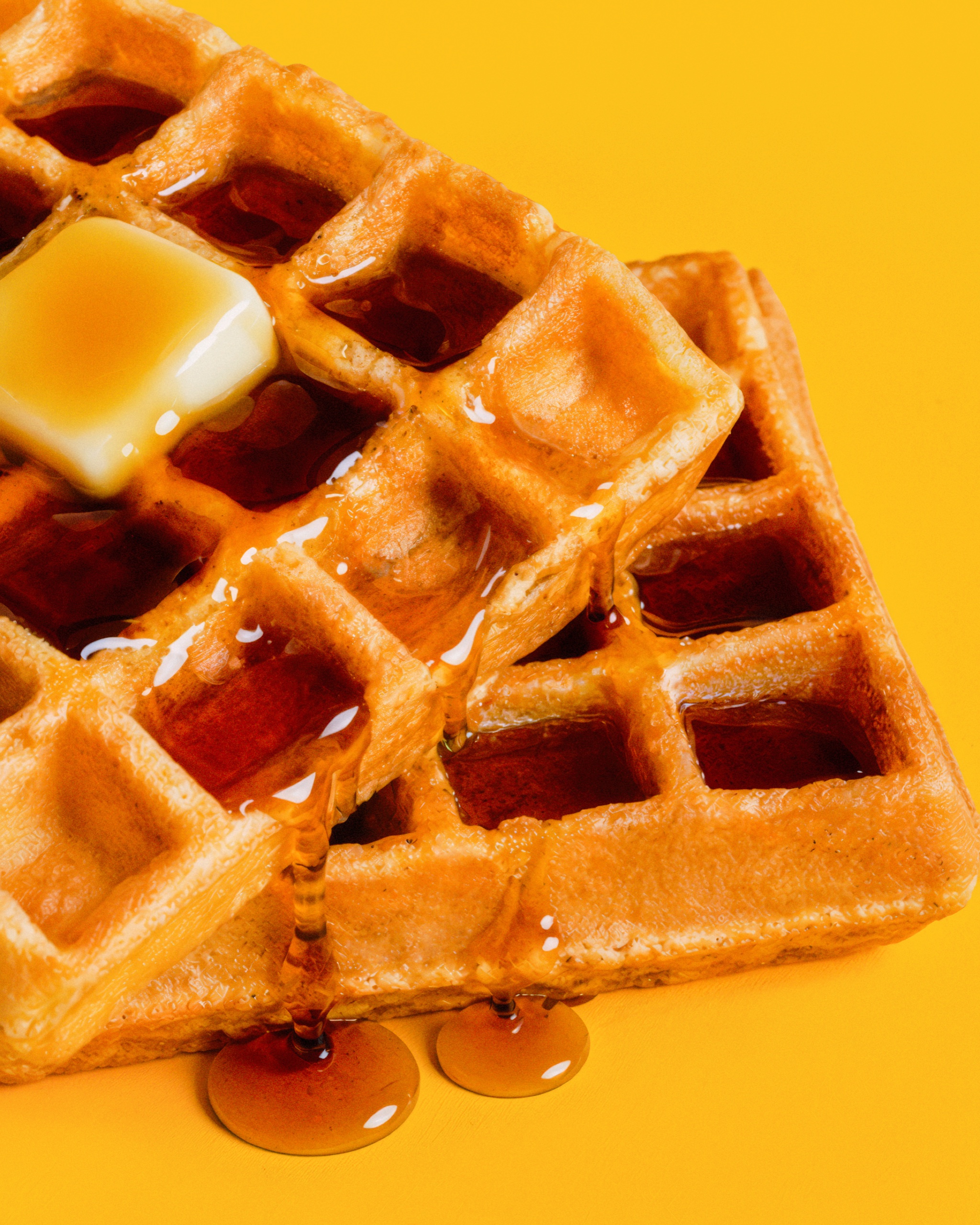 Close-up of golden waffles, melted butter, and syrup against yellow.