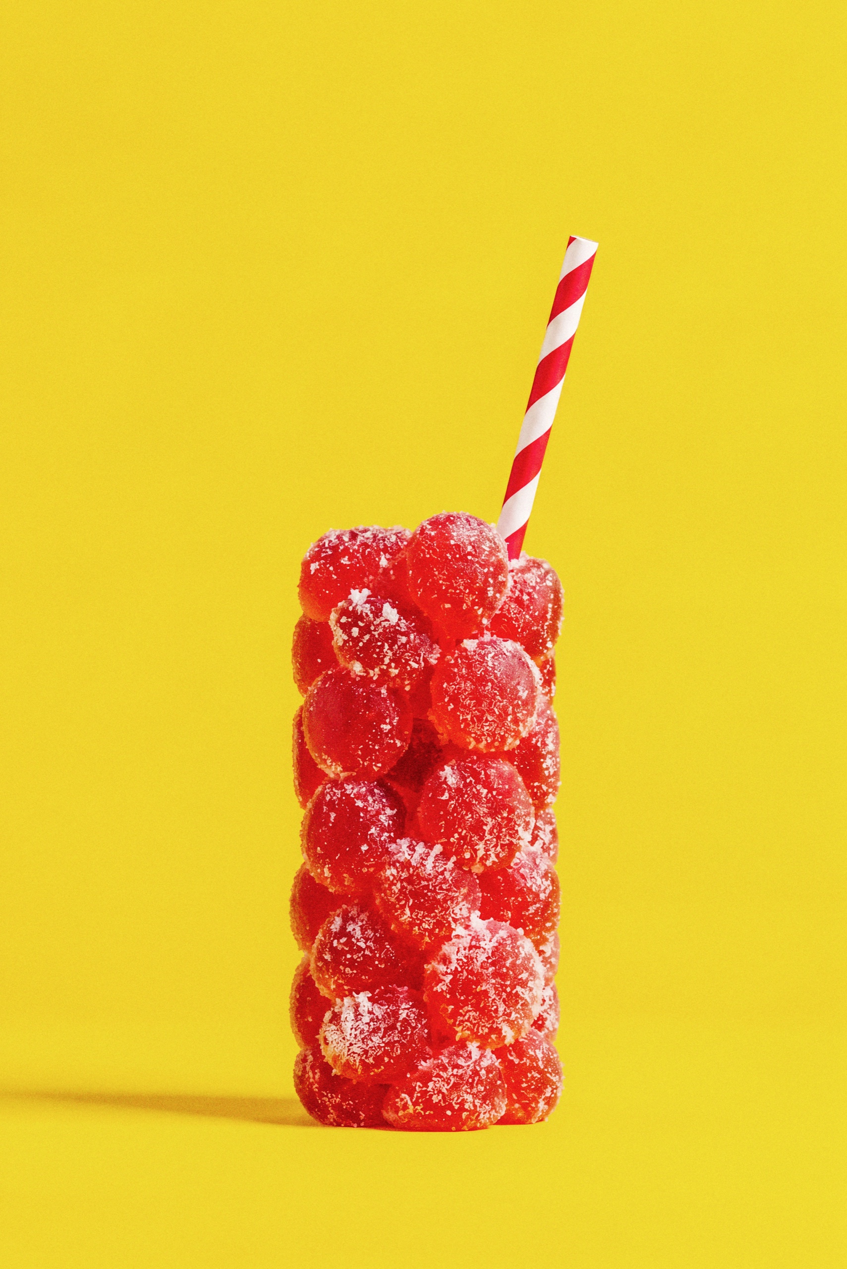 A tall drink-shaped stack of Collective Project Squeezies gummies with a red-and-white striped straw against yellow.