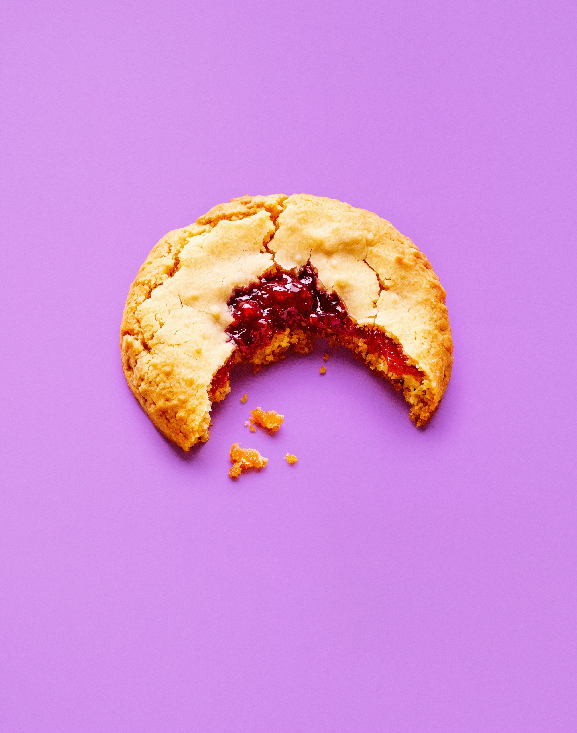 A golden cookie with red jam filling and a bite missing, with scattered crumbs against a purple background.