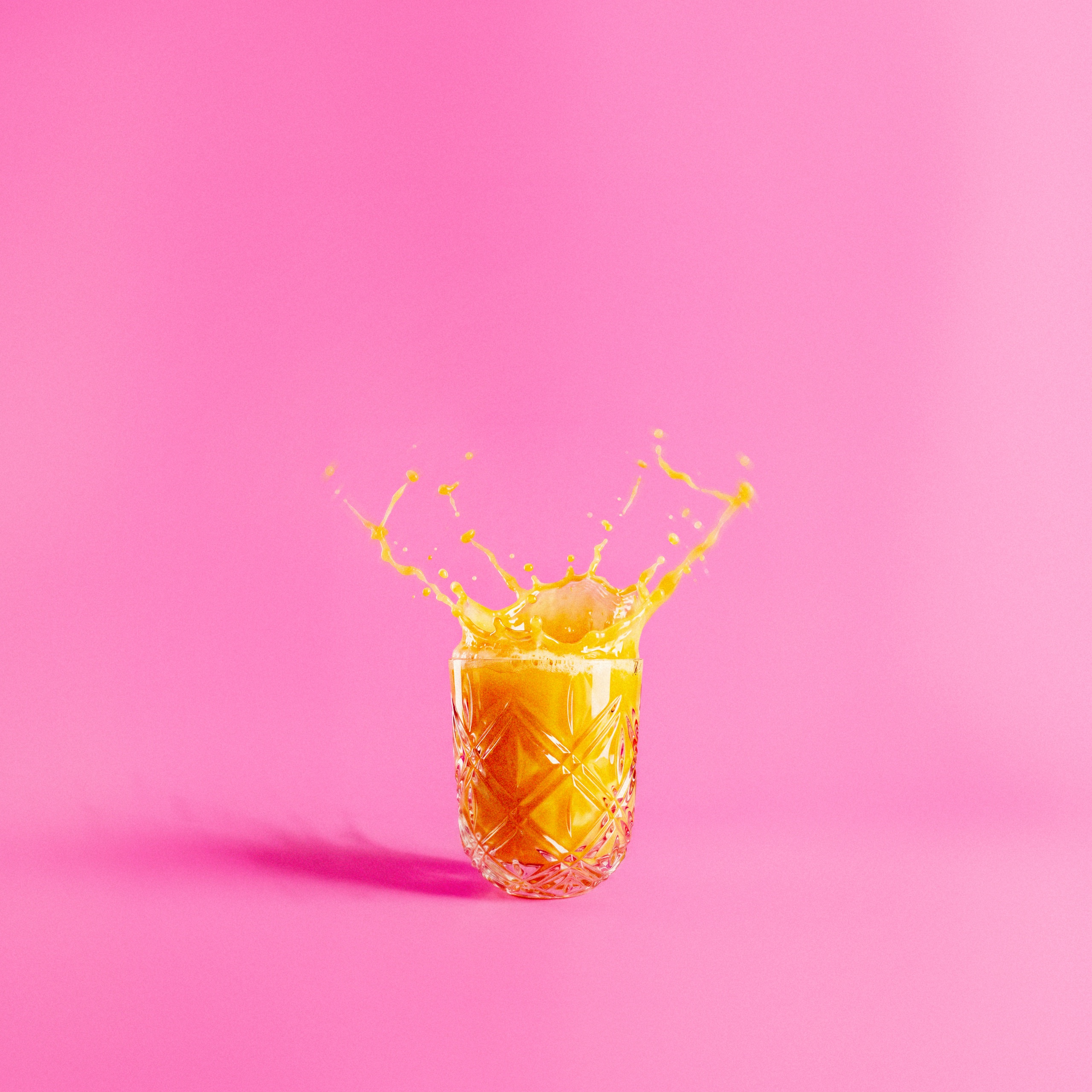 Orange juice splashes above a cut-glass tumbler on a vivid pink background.