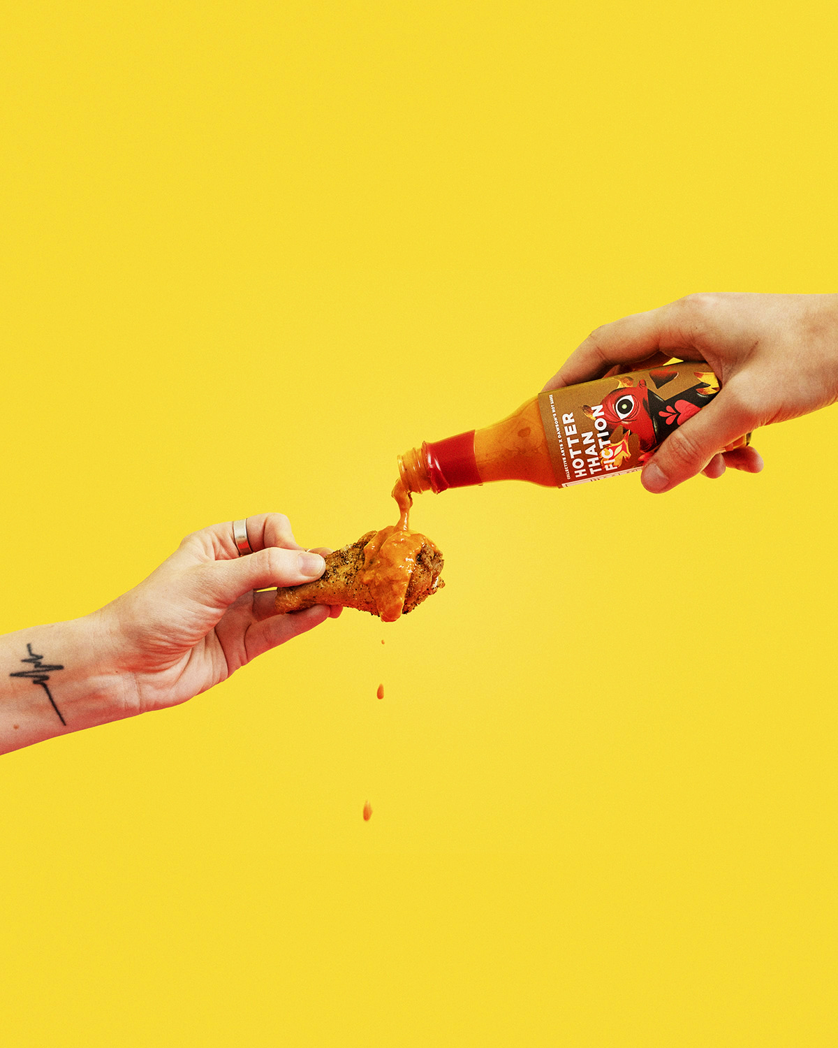 A hand pours Collective Arts and Dawson’s Hotter Than Fiction hot sauce onto a chicken wing against a yellow background.