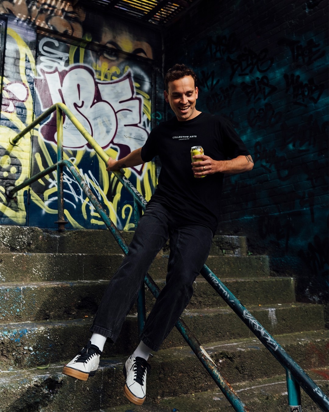 A man holding a Collective Arts beer slides down a handrail on graffiti-lined outdoor steps.