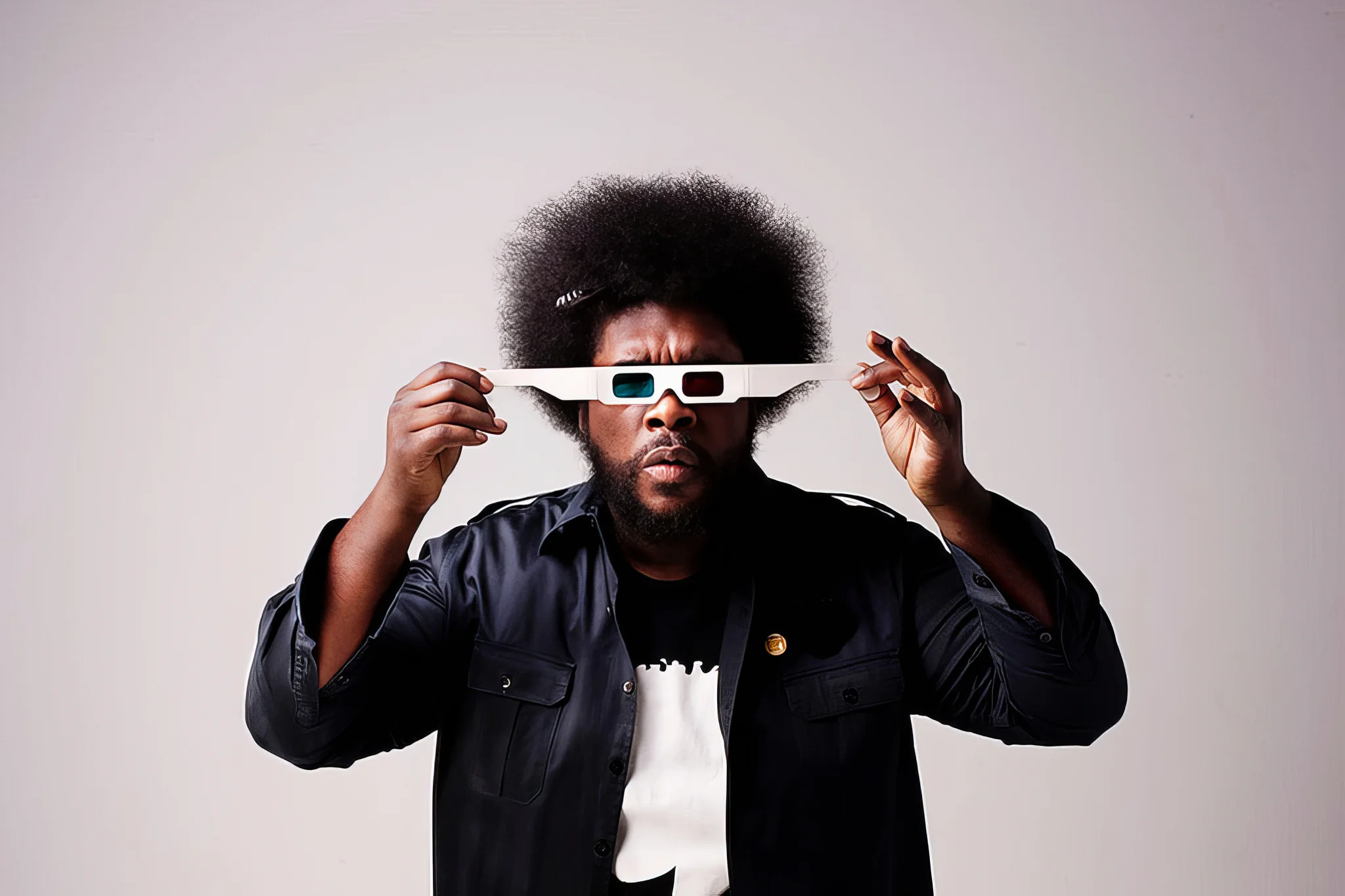 Colour studio portrait of a man with an afro holding oversized white 3D glasses.
