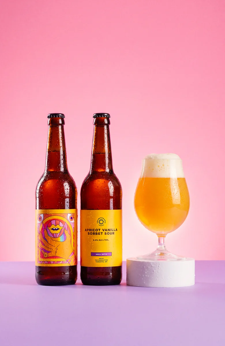 Two Collective Arts Apricot Vanilla Sorbet Sour bottles beside a glass of golden beer on a white pedestal, against a pink and purple backdrop.