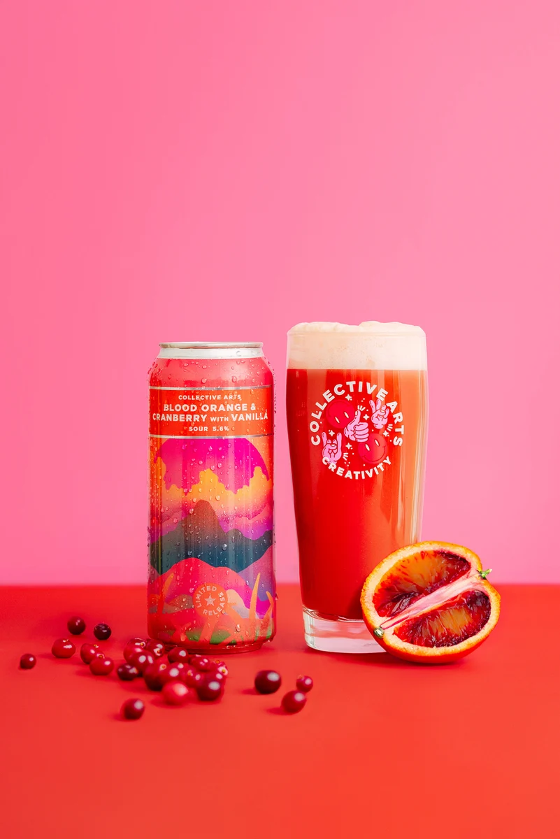 Collective Arts Blood Orange and Cranberry with Vanilla Sour can and glass, with cranberries and a halved blood orange on a red surface against pink.