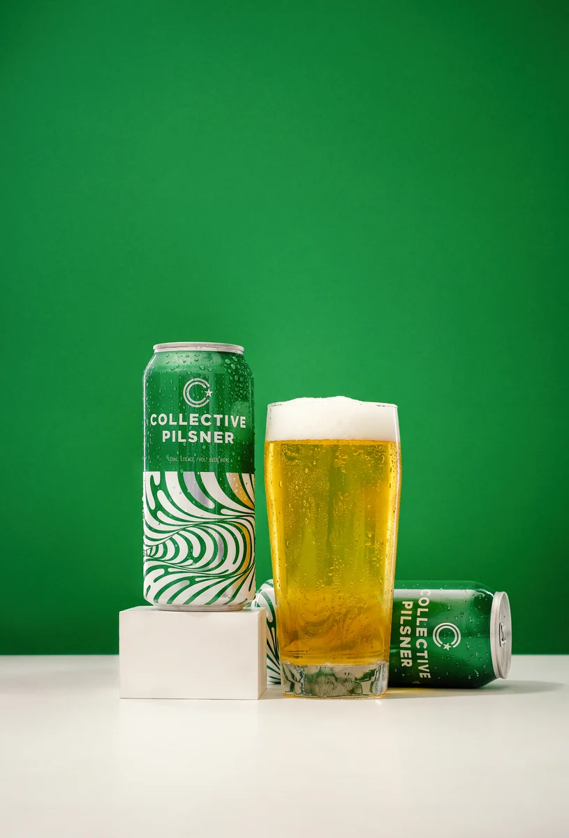 Two green Collective Pilsner cans beside a glass of golden beer, arranged on a white surface and pedestal against a green backdrop.