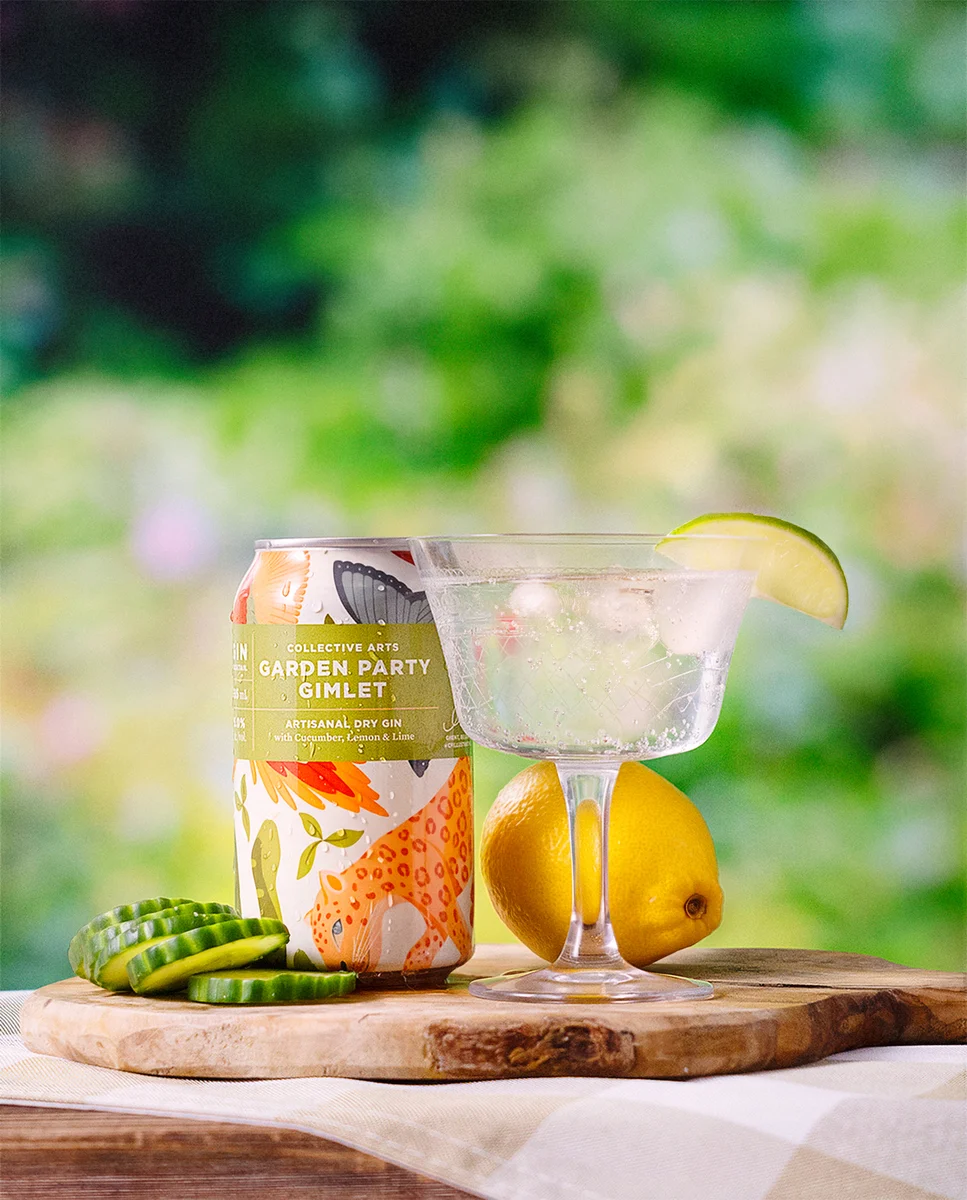 Collective Arts Garden Party Gimlet can and cocktail with lime, lemon and cucumber on a wooden board against a leafy green background.