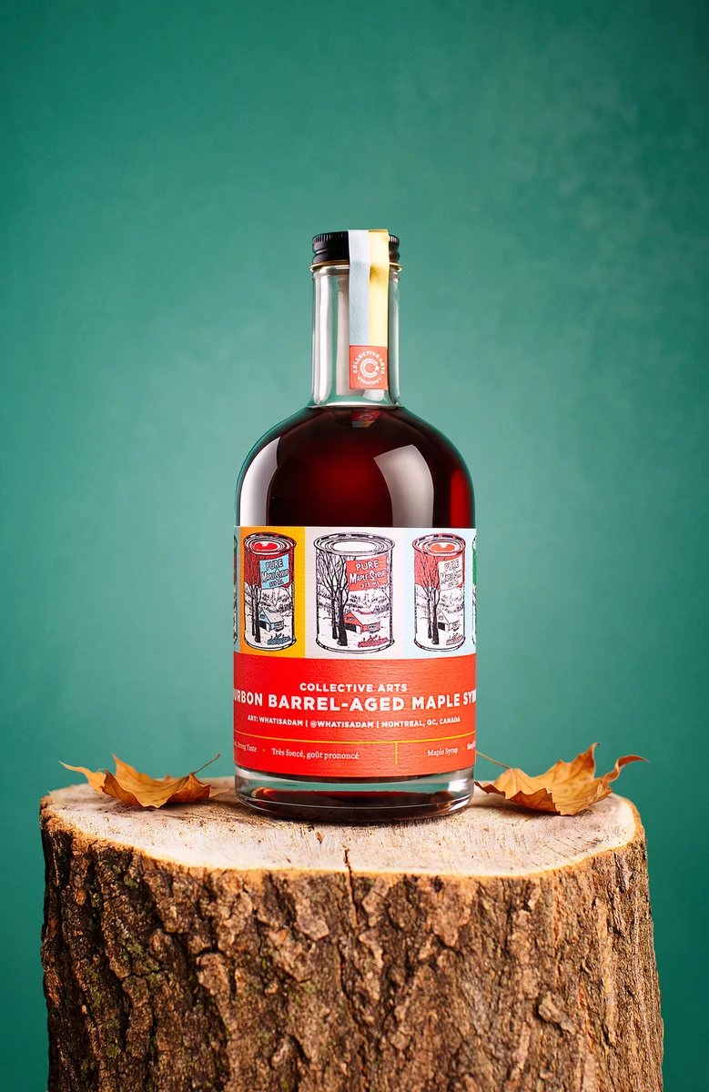 Collective Arts bourbon barrel-aged maple syrup bottle on a tree stump with autumn leaves against a green background.