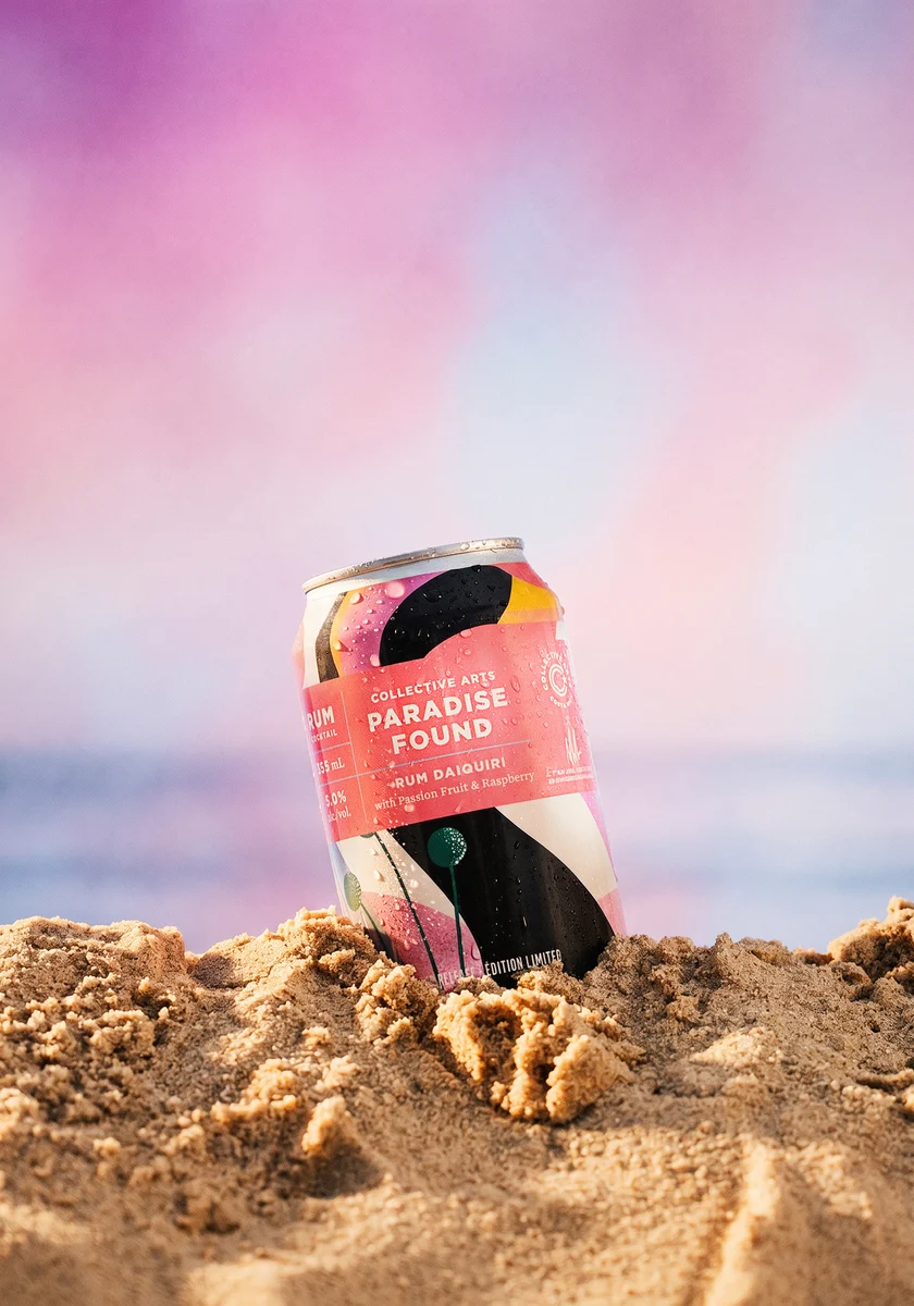 Collective Arts Paradise Found rum daiquiri can nestled in sand against a soft pink and blue background.
