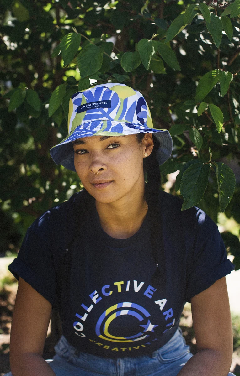 Portrait of a model wearing a blue and yellow patterned Collective Arts bucket hat among leafy branches.