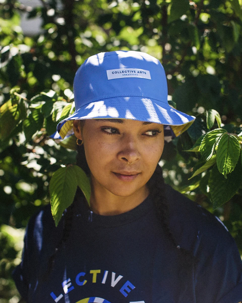 Close-up of a model wearing a blue Collective Arts bucket hat in dappled sunlight.