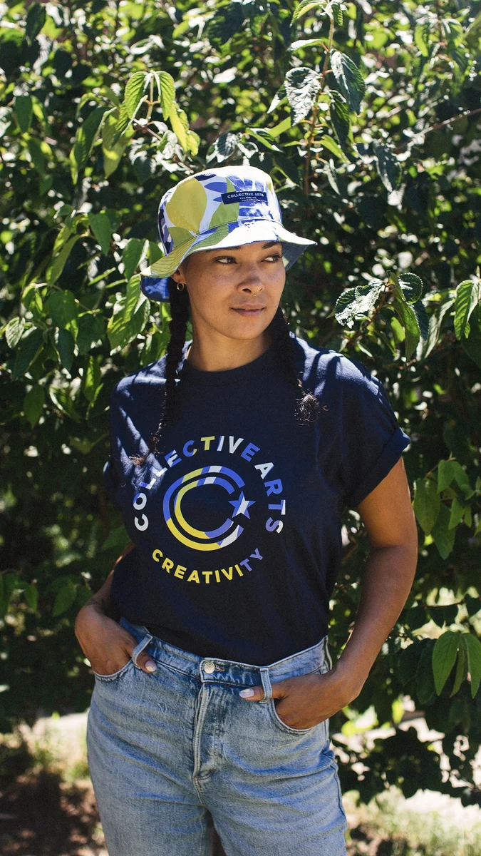 Model looking to the side, wearing a patterned Collective Arts bucket hat and navy graphic T-shirt in front of green foliage.