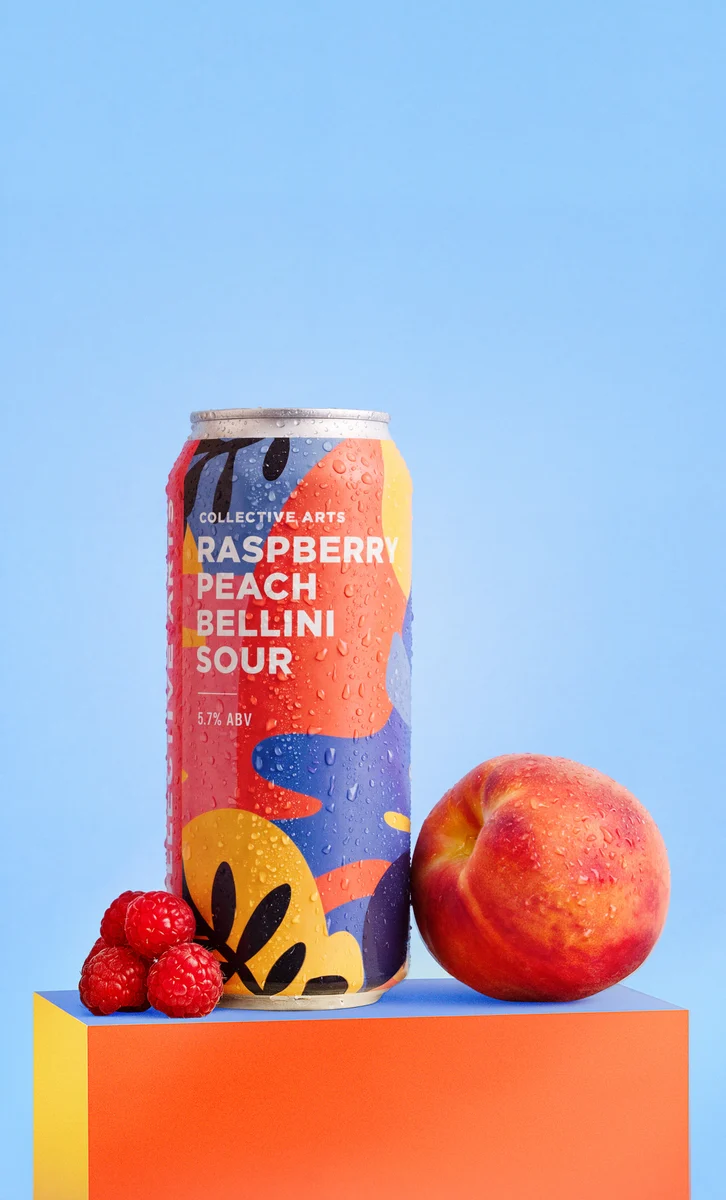 Collective Arts Raspberry Peach Bellini Sour can beside a peach and raspberries on a colourful pedestal against blue.