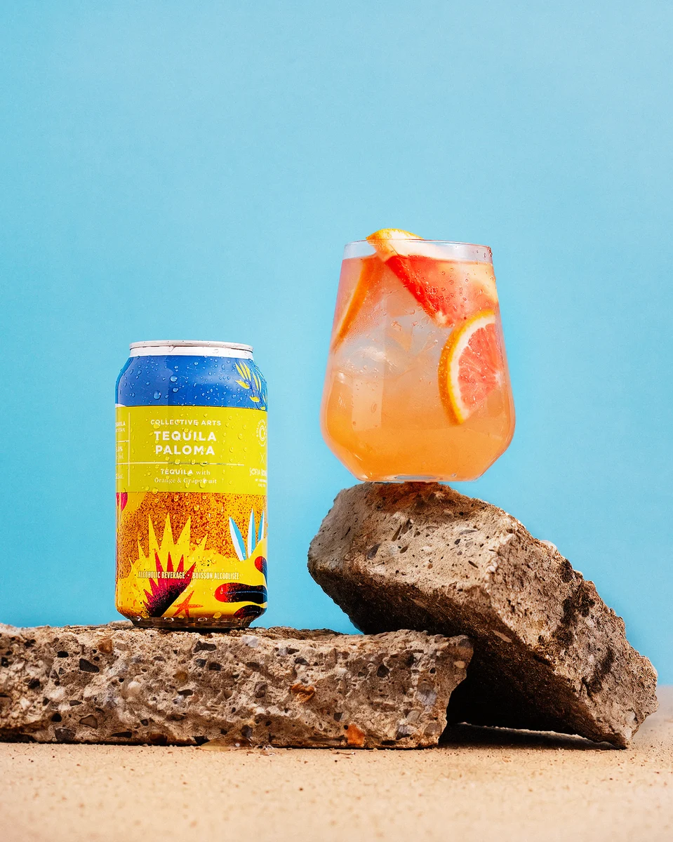 Collective Arts Tequila Paloma can and citrus-garnished cocktail arranged on rocks against a blue background.