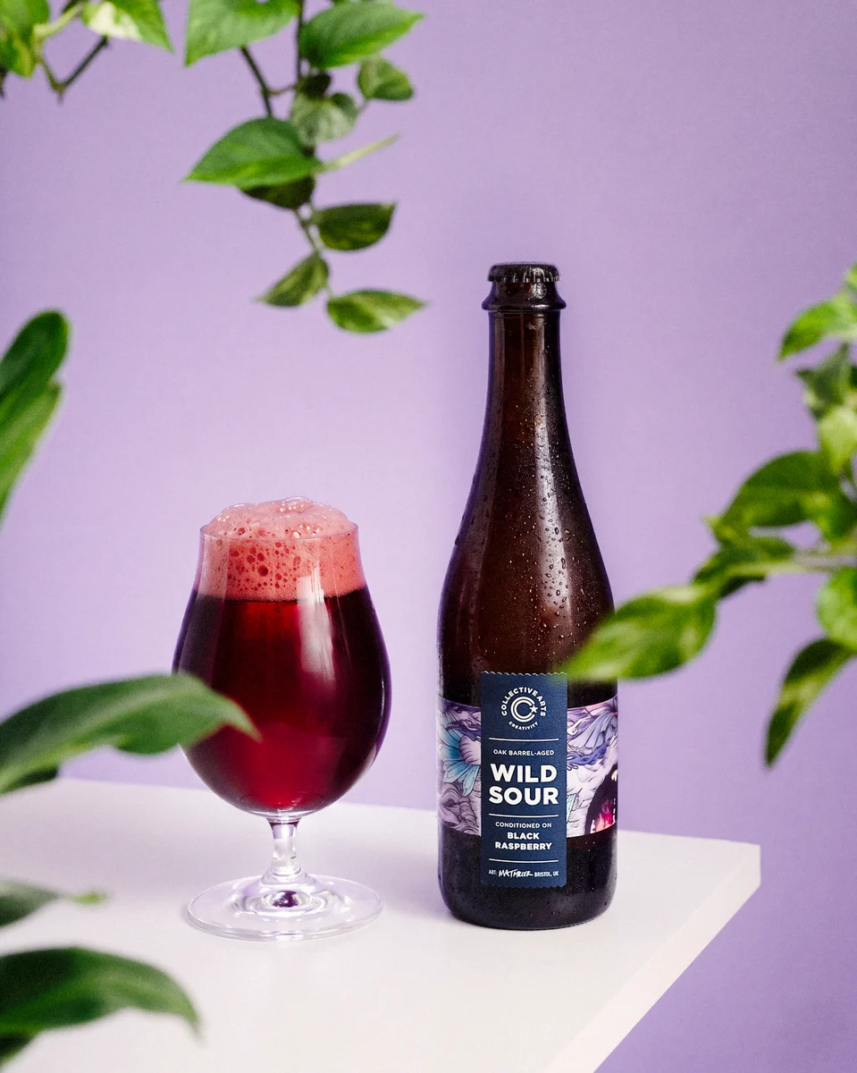 Collective Arts Black Raspberry Wild Sour bottle and a glass of deep red beer, framed by green leaves against a purple backdrop.